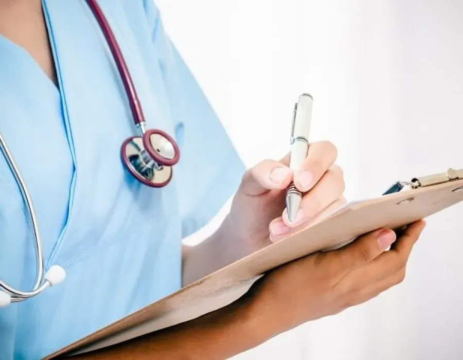 Practical nurse in blue scrubs and wearing stethoscope taking notes on clipboard